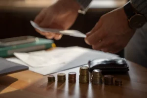 Mãos manuseando documentos ou notas sobre uma mesa, com pilhas de moedas e uma carteira em primeiro plano. A cena representa a organização de finanças e questões patrimoniais.
