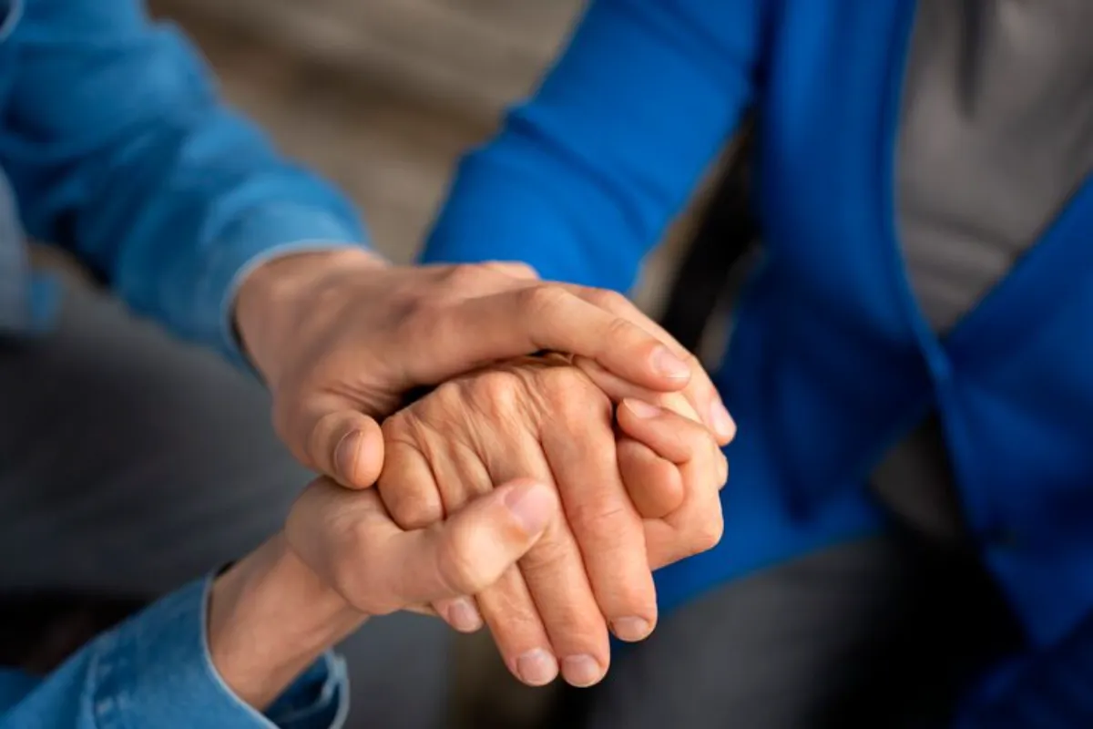 Duas pessoas de mãos dadas, com uma mão mais jovem envolvendo a mão de uma pessoa idosa, simbolizando apoio emocional durante o luto.