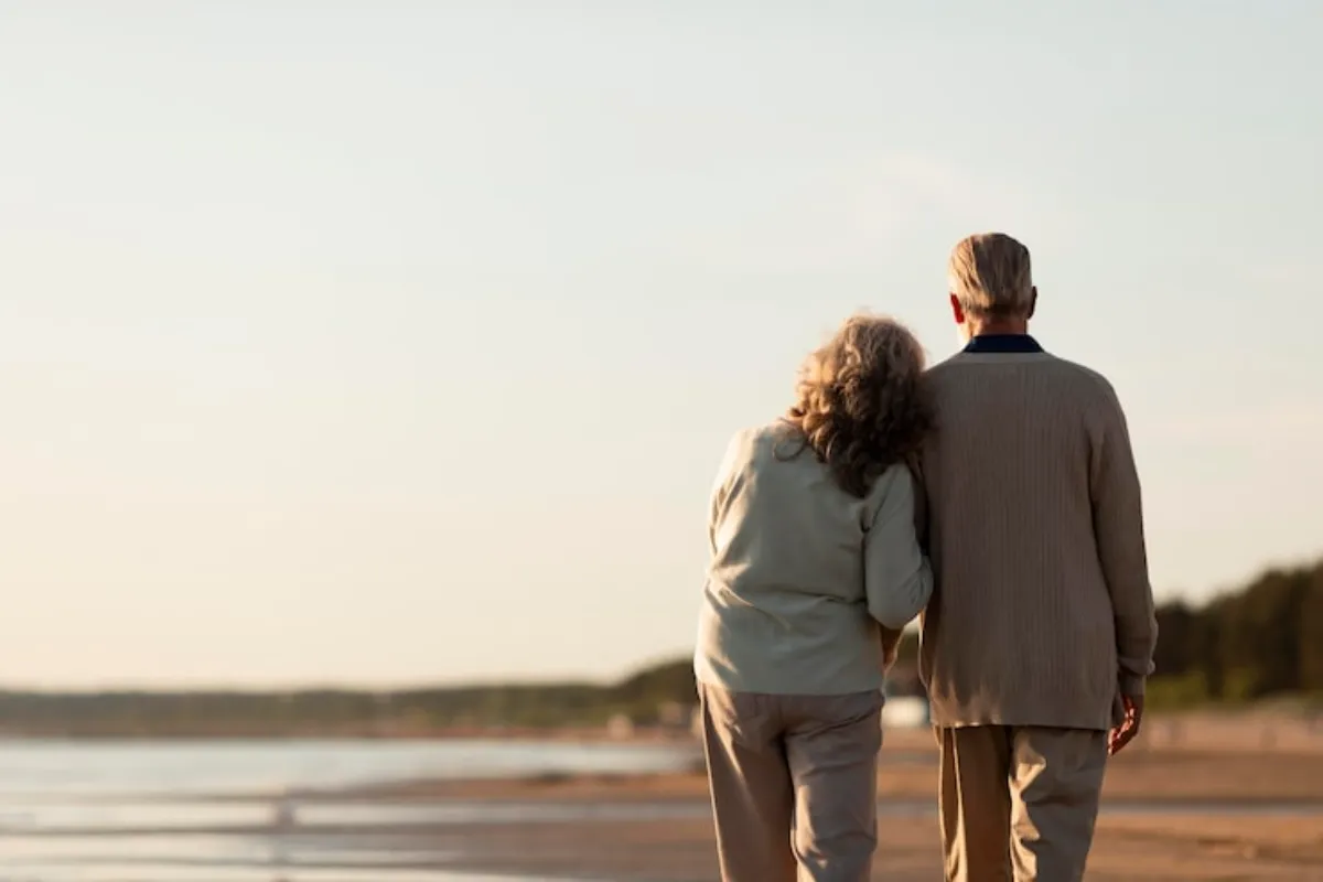 Casal de idosos caminhando juntos pela praia ao entardecer.